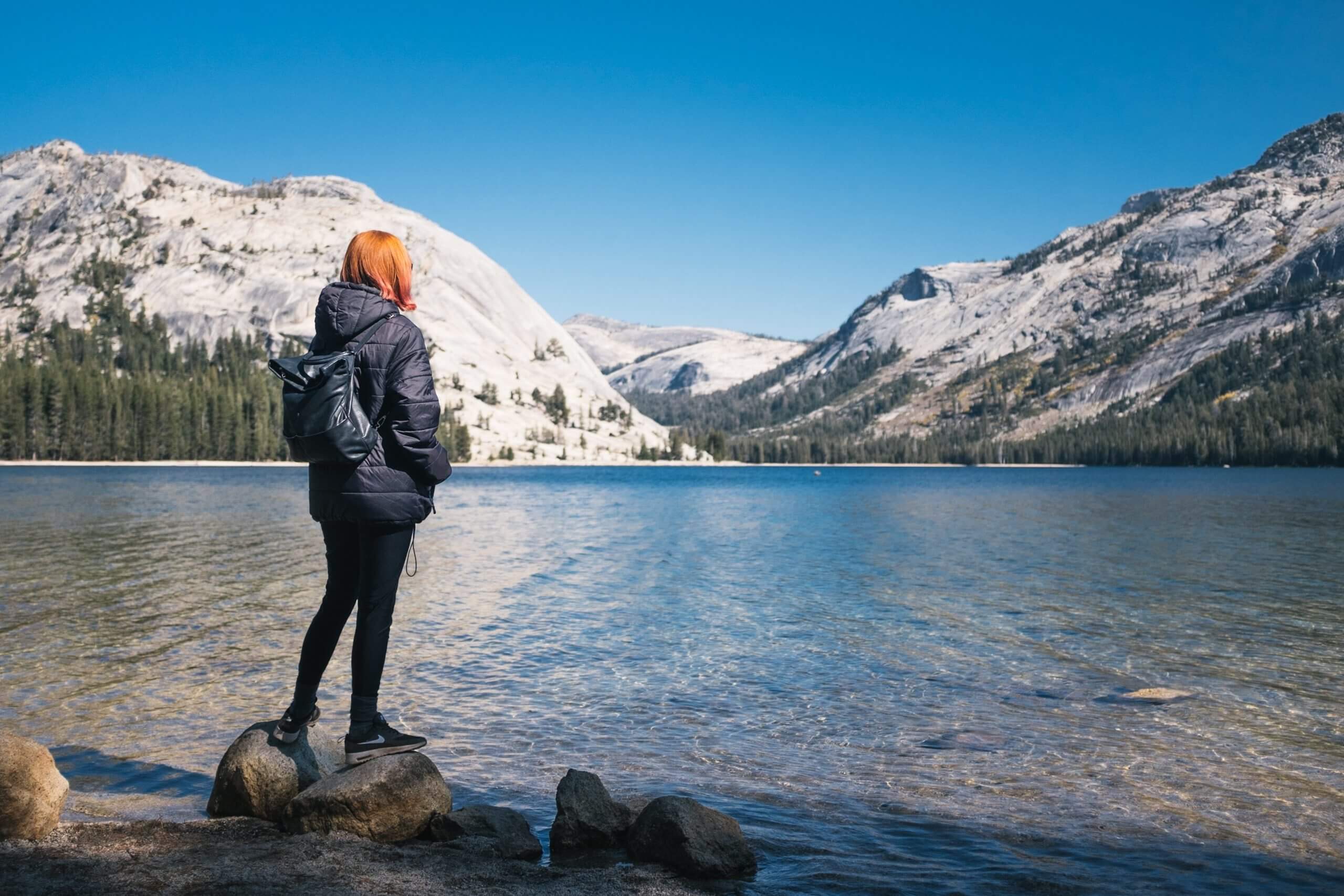 Red haired person wearing coat and carrying backpack surveys a lake and distant snow-covered mountain