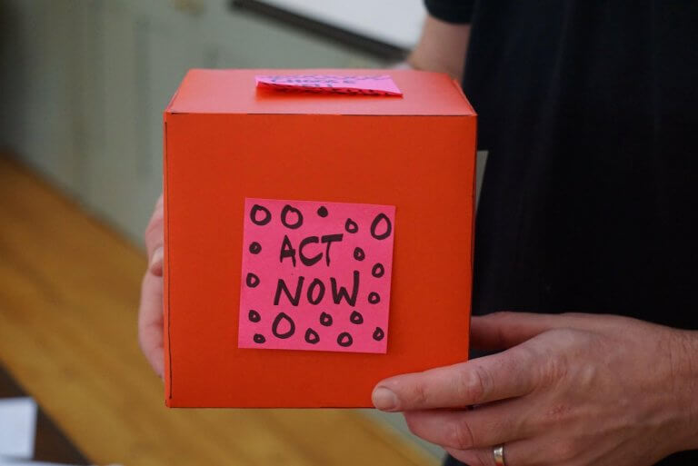 Person holding red box with a pink post-it note on the side that reads "Act Now"