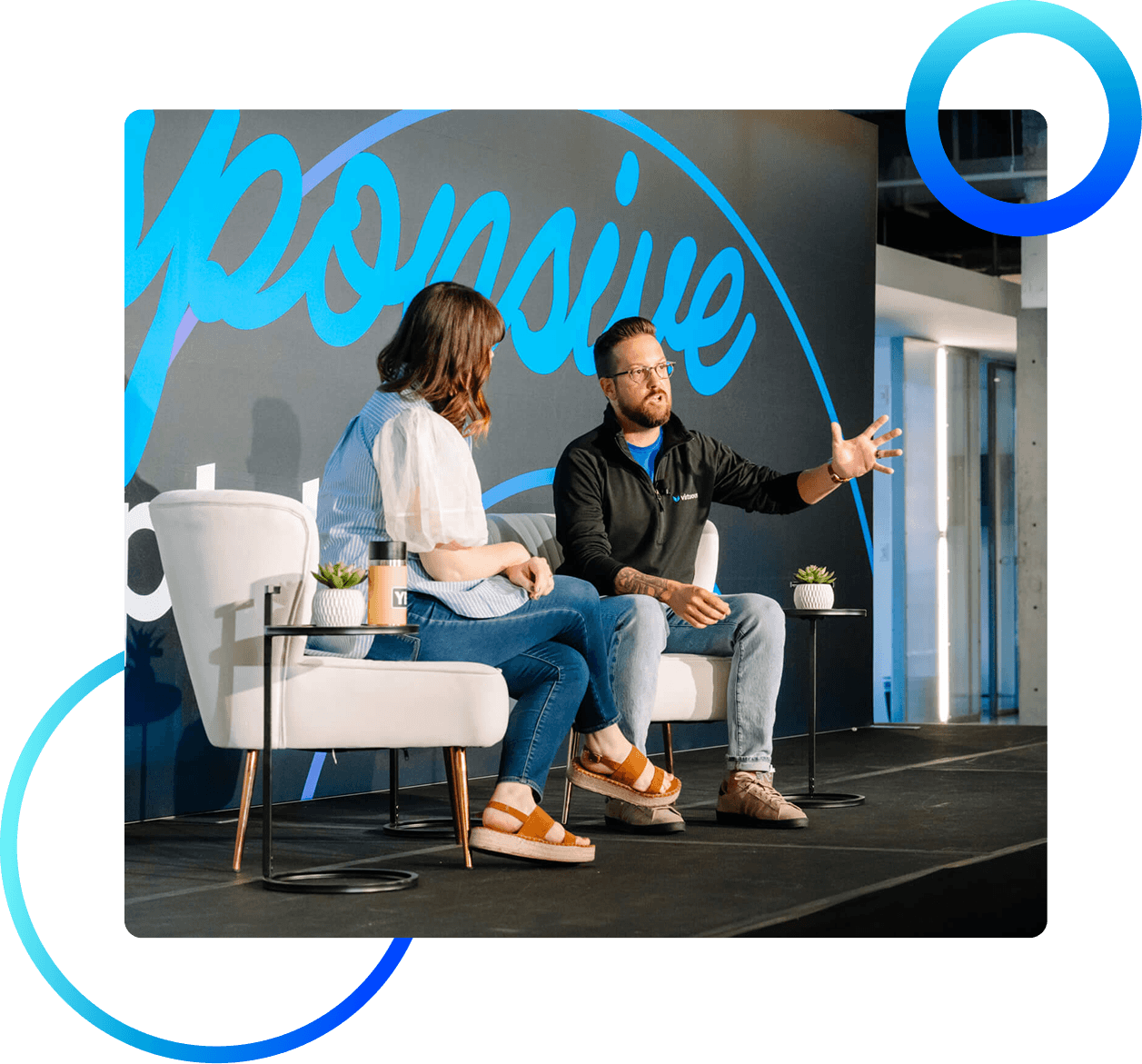 Two people on a stage in a panel discussion, with a large blue mural behind them and small side tables with plants nearby.